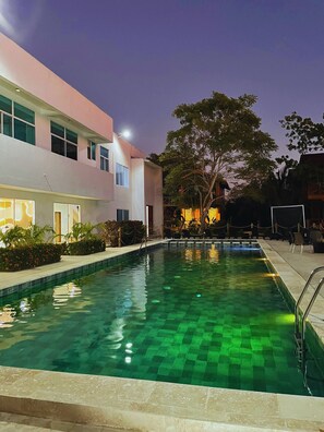 Una piscina con solárium de baldosas, rodeada por un edificio con ventanales grandes y un árbol.