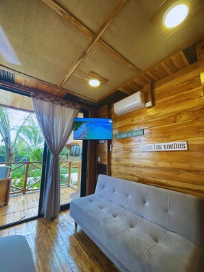 Una habitación con techo de madera, un sofá gris, un televisor montado en la pared y vistas a un balcón con plantas.