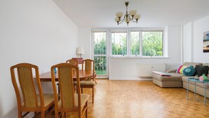 Een kamer met een eettafel en stoelen, een bank, een radiator en een raam met uitzicht op groen.