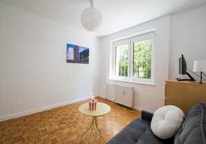 A modern living room with a grey sofa, a small round table, and a radiator. There is a window with a view of greenery outside.
