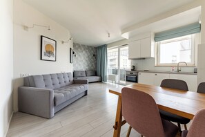 Eine moderne Wohnung mit einem grauen Sofa, einem Esstisch mit Stühlen, einer Küchenzeile und einem großen Fenster.