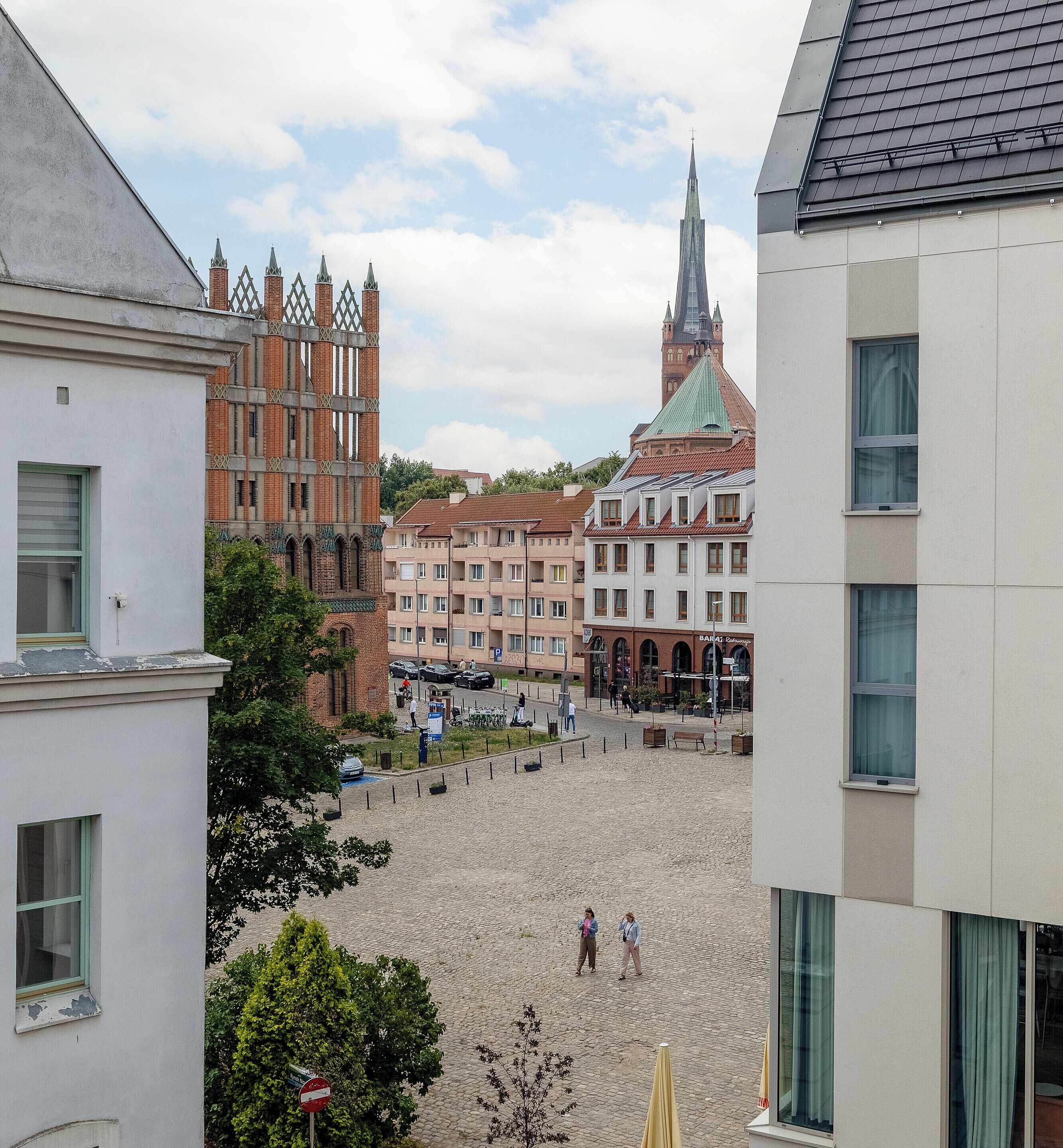 A cobblestone square with historic buildings, a church spire, and a modern building with a flat roof.