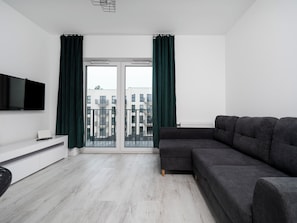 A modern living room with a grey sofa, a flat-screen TV mounted on the wall, and a view of the city through large windows with green curtains.