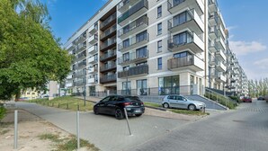 Kerrostalo, jossa on parvekkeita, asfaltoitu jalankulkualue ja parkkeerattuja autoja.