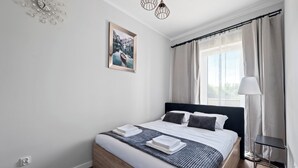 A modern bedroom with a bed, a bedside table, a lamp, and a window with curtains. - Pagi City Comfort Apartment with Garden (Lublin)