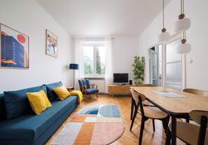 Una sala de estar moderna con un sofá azul, cojines amarillos, una mesa de comedor de madera y un televisor.