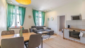Ein modernes Wohnzimmer mit grauem Sofa, einem Holzespeisetisch und Stühlen. Es gibt eine Küchenzeile mit Mikrowelle, Backofen und einem über der Arbeitsfläche montierten Fernseher.