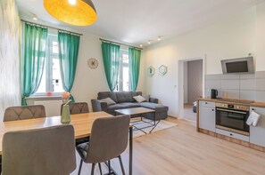 A modern living room with a grey sofa, a wooden dining table, and chairs. There is a kitchen area with a microwave, oven, and a TV mounted above the counter. - Mikołowska Silesian Charm Apartment (Katowice)