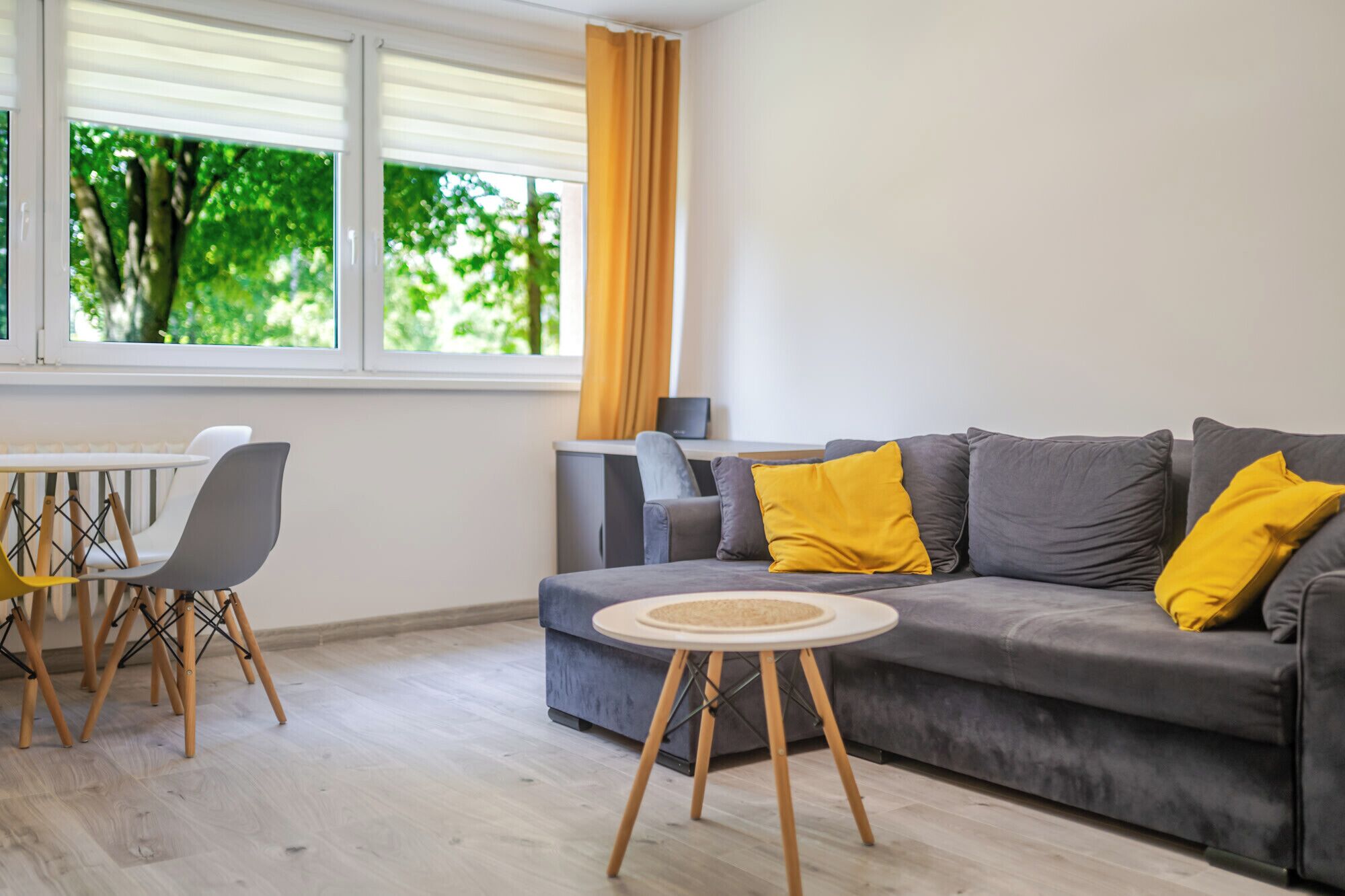 Una sala moderna con un sofá gris, cojines amarillos, una mesa de centro redonda y un comedor con mesa y sillas blancas.