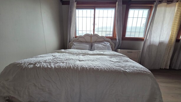 Een groot bed met een witte spreidekbed in een kamer met houten vloeren en twee ramen met gordijnen.