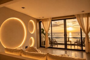 A modern interior with a large window overlooking the ocean at sunset, a seating area with cushions, and a wall with illuminated circular designs. - Water-front Luxury + King Bed w/AC (Tijuana)