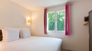 Hotellihuone, jossa on sänky, ikkuna verhoilla, seinään kiinnitetty valaisin ja vaatekaappi.