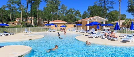 Piscina con sillones de descanso y sombrillas, rodeada de árboles y edificios.
