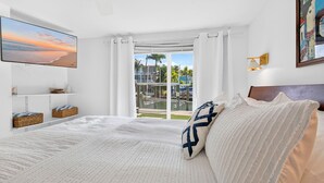 Ein Schlafzimmer mit einem Bett, einem Fernseher, einem Fenster mit Vorhängen, einem Gemälde und Blick auf einen Pool sowie Gebäude.