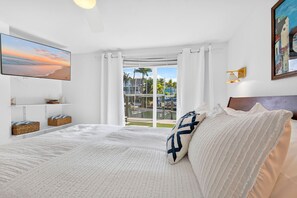 A bedroom with a bed, a TV, a window with curtains, a painting, and a view of a pool and buildings. - Pet Friendly Canal Home - Boat Lift + Paddleboards (Sarasota)