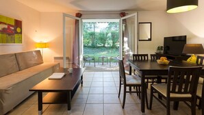 A living room with a sofa, coffee table, and dining table with chairs. There is a door leading to a garden. - Piscines & tennis sur place | Terrasse privée (Barbaste)