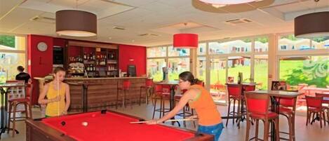 A pool table in a brightly lit indoor space with a bar area and red chairs.
