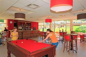 A pool table in a brightly lit indoor space with a bar area and red chairs.