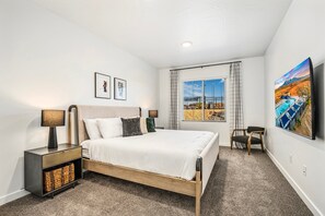Une chambre à coucher avec un lit, des tables de chevet, une fenêtre avec des rideaux, une chaise et des œuvres d’art accrochées au mur.