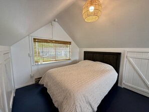 Een slaapkamer met een bed, een raam met zonwering, een hanglamp en een schuine plafond.