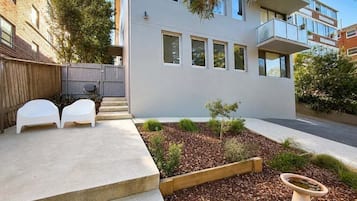 A modern building with a flat roof, large windows, and a small garden area with a wooden planter and two white chairs.