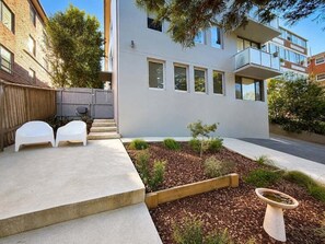 A modern building with a flat roof, large windows, and a small garden area with a wooden planter and two white chairs.