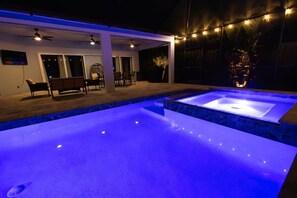 An outdoor swimming pool with a jacuzzi, surrounded by a covered patio with seating and string lights. - Modern Oasis (Sarasota)