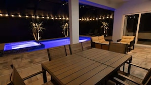 An outdoor patio with a dining table and chairs, a small pool with blue lighting, and string lights. - Modern Oasis (Sarasota)