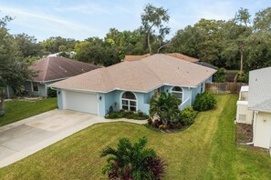 A residential house with a light blue exterior, a brown roof, and a front yard with grass and small plants. - Modern Oasis (Sarasota)