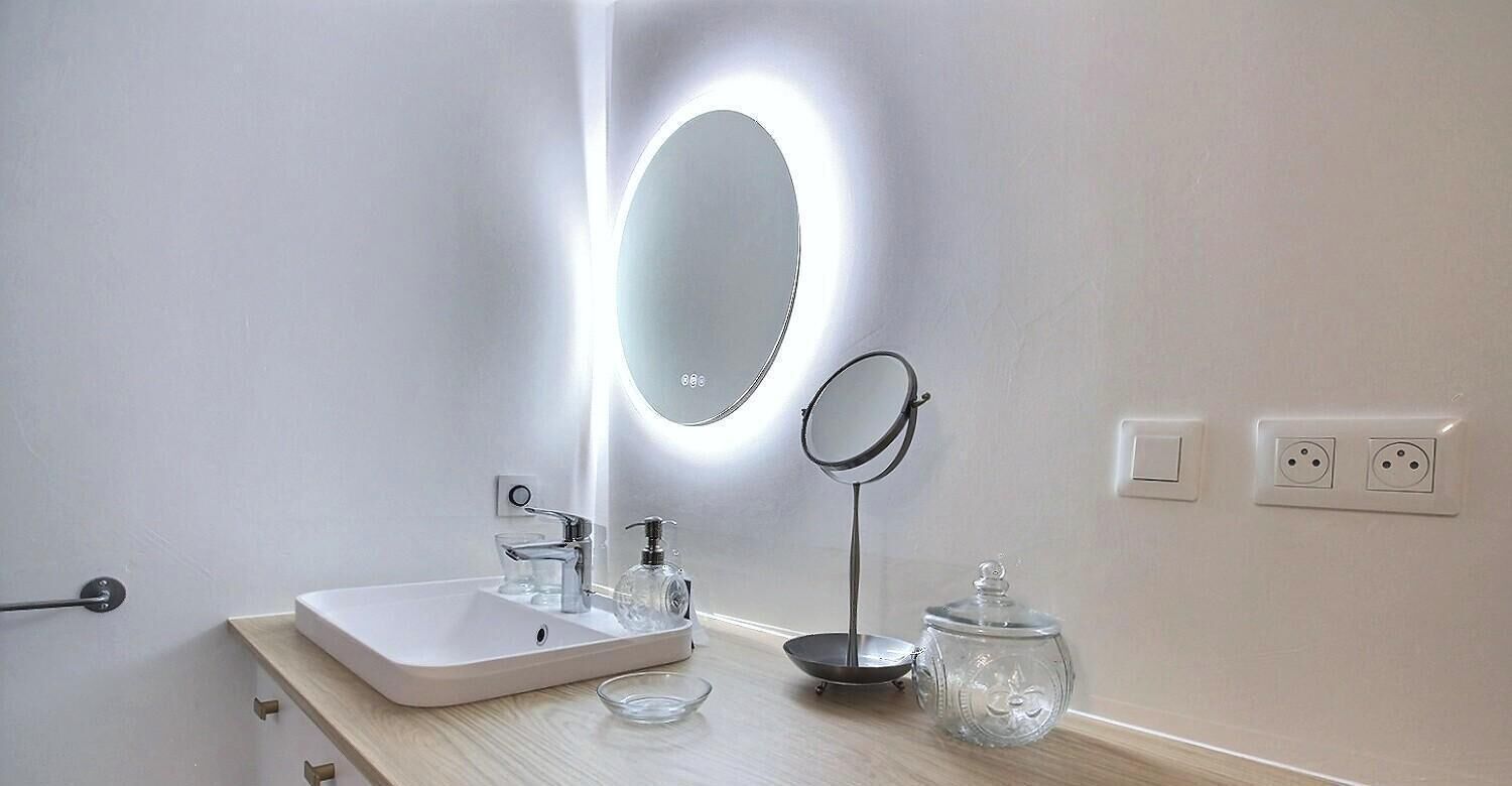 A bathroom with a white sink, a round mirror, a small round mirror on a stand, a glass jar, and a light above the mirror.