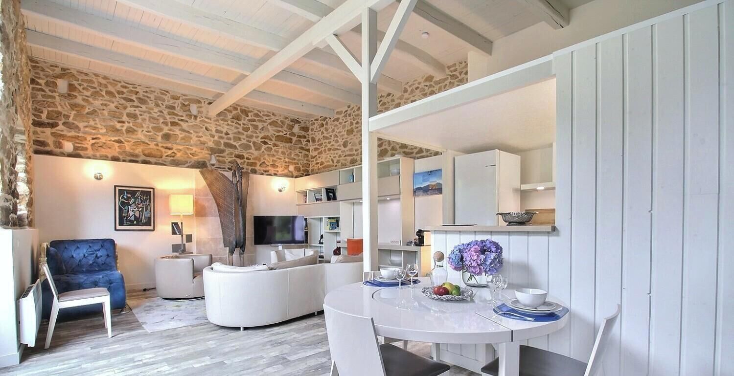 A modern living room with a stone wall, a blue armchair, a white sofa, a television, and a dining area with a table set for two.