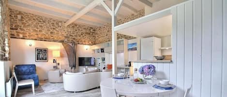 A modern living room with a stone wall, a blue armchair, a white sofa, a television, and a dining area with a table set for two.