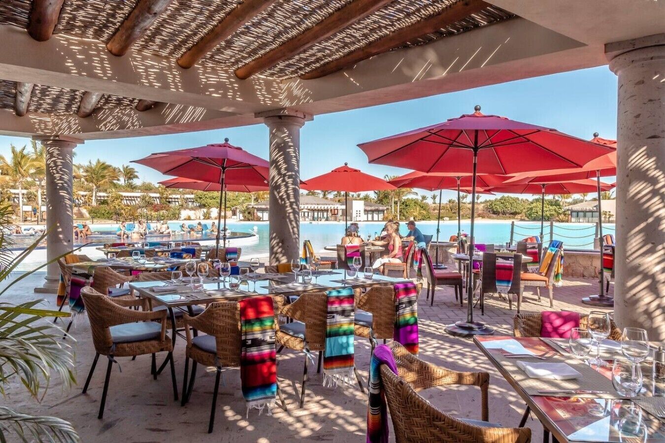 An outdoor dining area with tables and chairs, red umbrellas, and a pool in the background.