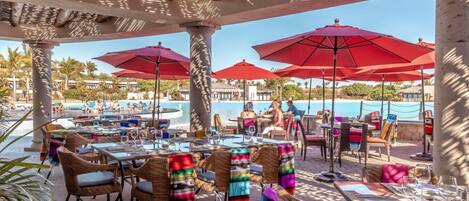 An outdoor dining area with tables and chairs, red umbrellas, and a pool in the background.
