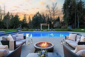 A swimming pool with lounge chairs and a fire pit in the center, surrounded by trees and a gazebo.
