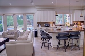 A modern kitchen with white cabinets, a central island with bar stools, and large windows offering a view of the outdoors. - Luxury Mansion Pickleball court, Sleeps 26 Pool/Spa/Playground/Theatre/Game Room (Langley Township)