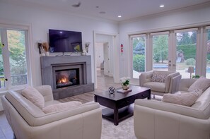 A modern living room with a fireplace, comfortable seating, a flat-screen TV, and large windows. - Luxury Mansion Pickleball court, Sleeps 26 Pool/Spa/Playground/Theatre/Game Room (Langley Township)