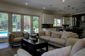 A spacious living area with a sofa, coffee table, and a view of a swimming pool through large windows. - Luxury Mansion Pickleball court, Sleeps 26 Pool/Spa/Playground/Theatre/Game Room (Langley Township)