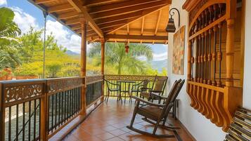 A covered balcony with wooden railings, a rocking chair, and a view of greenery and mountains.