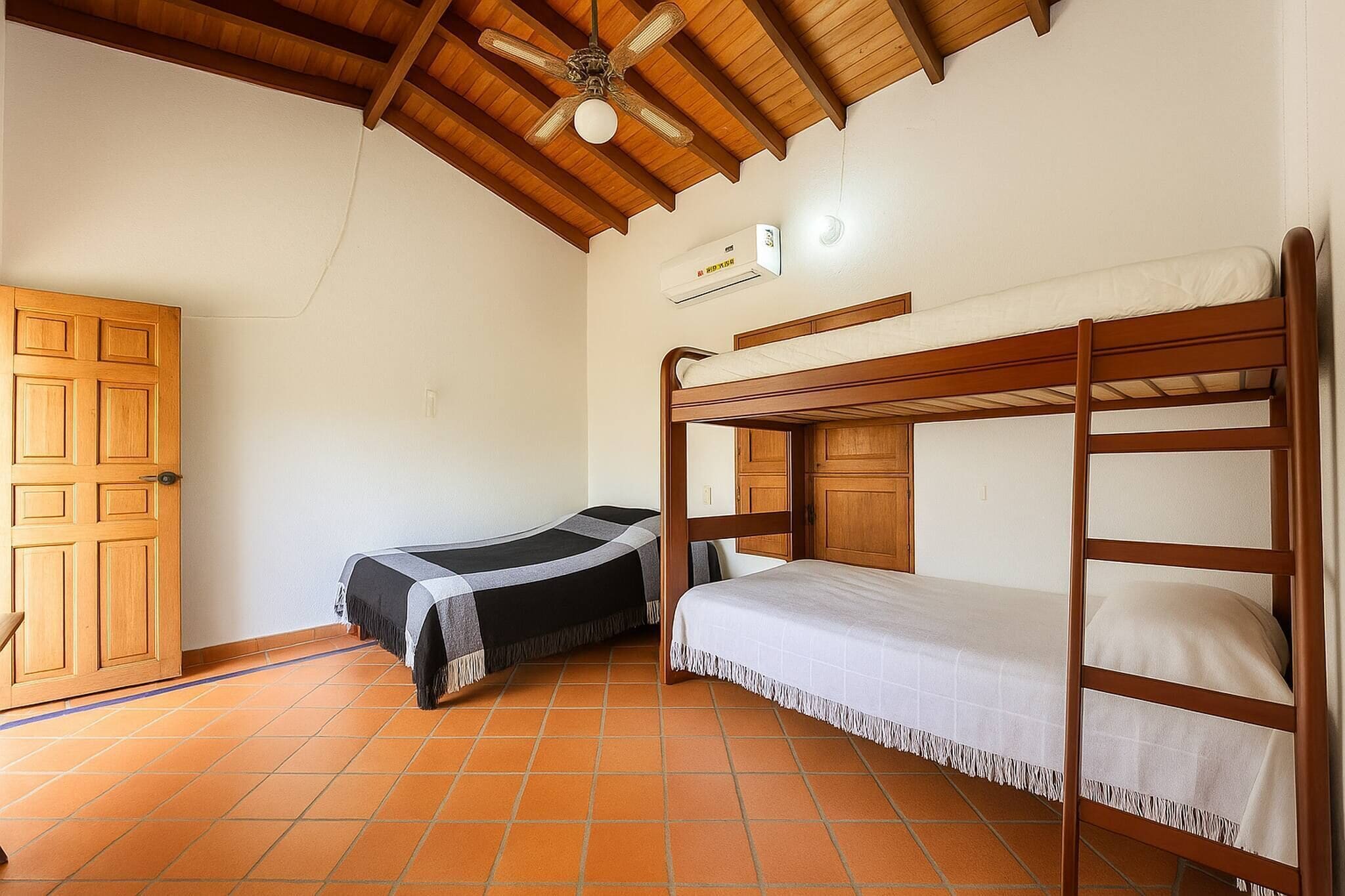 A room with a wooden ceiling, a bunk bed, a single bed, a wardrobe, and a tiled floor.