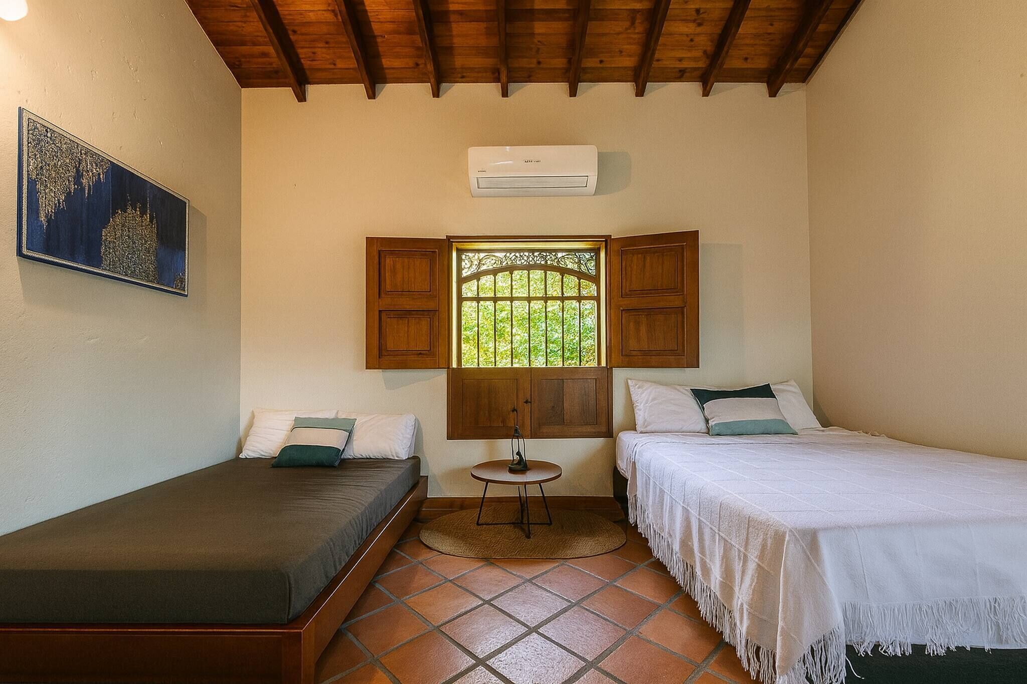 Una habitación pequeña con dos camas, una ventana con contraventanas de madera, una mesita y un equipo de aire acondicionado.