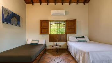 Una habitación pequeña con dos camas, una ventana con contraventanas de madera, una mesita y un equipo de aire acondicionado.
