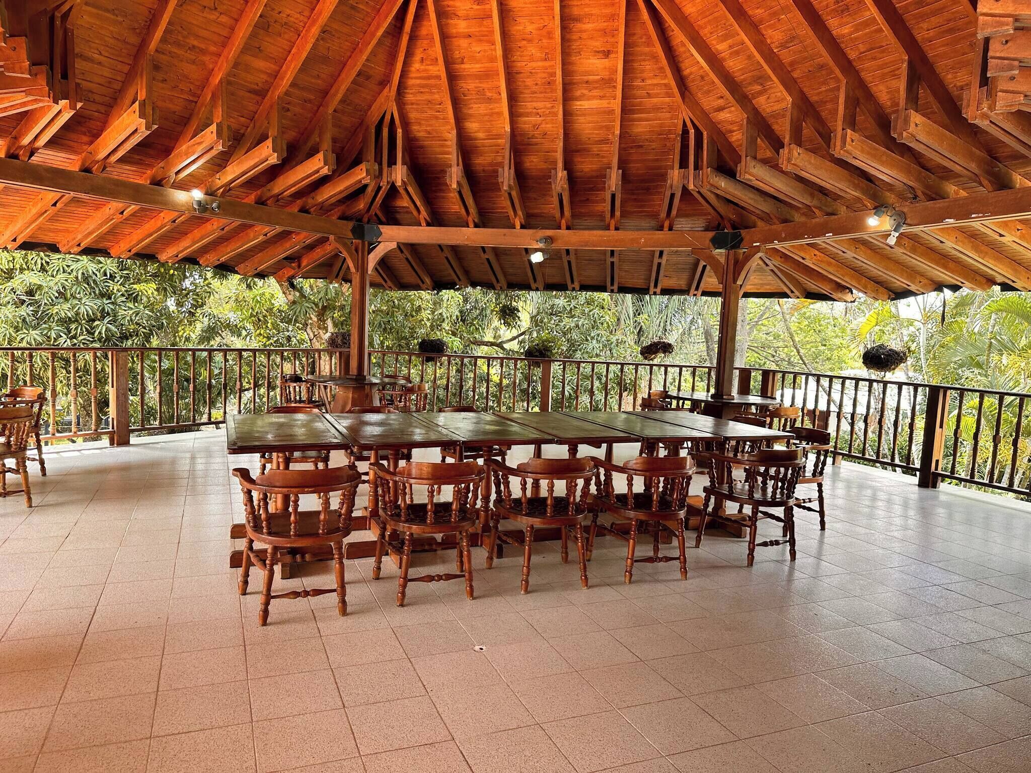 Un área de comedor al aire libre, cubierta, con mesas y sillas de madera, rodeada por una barandilla y árboles.