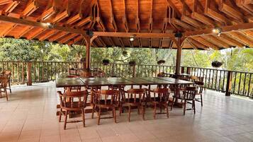 Un área de comedor al aire libre, cubierta, con mesas y sillas de madera, rodeada por una barandilla y árboles.