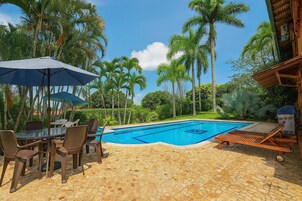 Un área de piscina con mesa y sillas, rodeada de palmeras y una estructura de madera.
