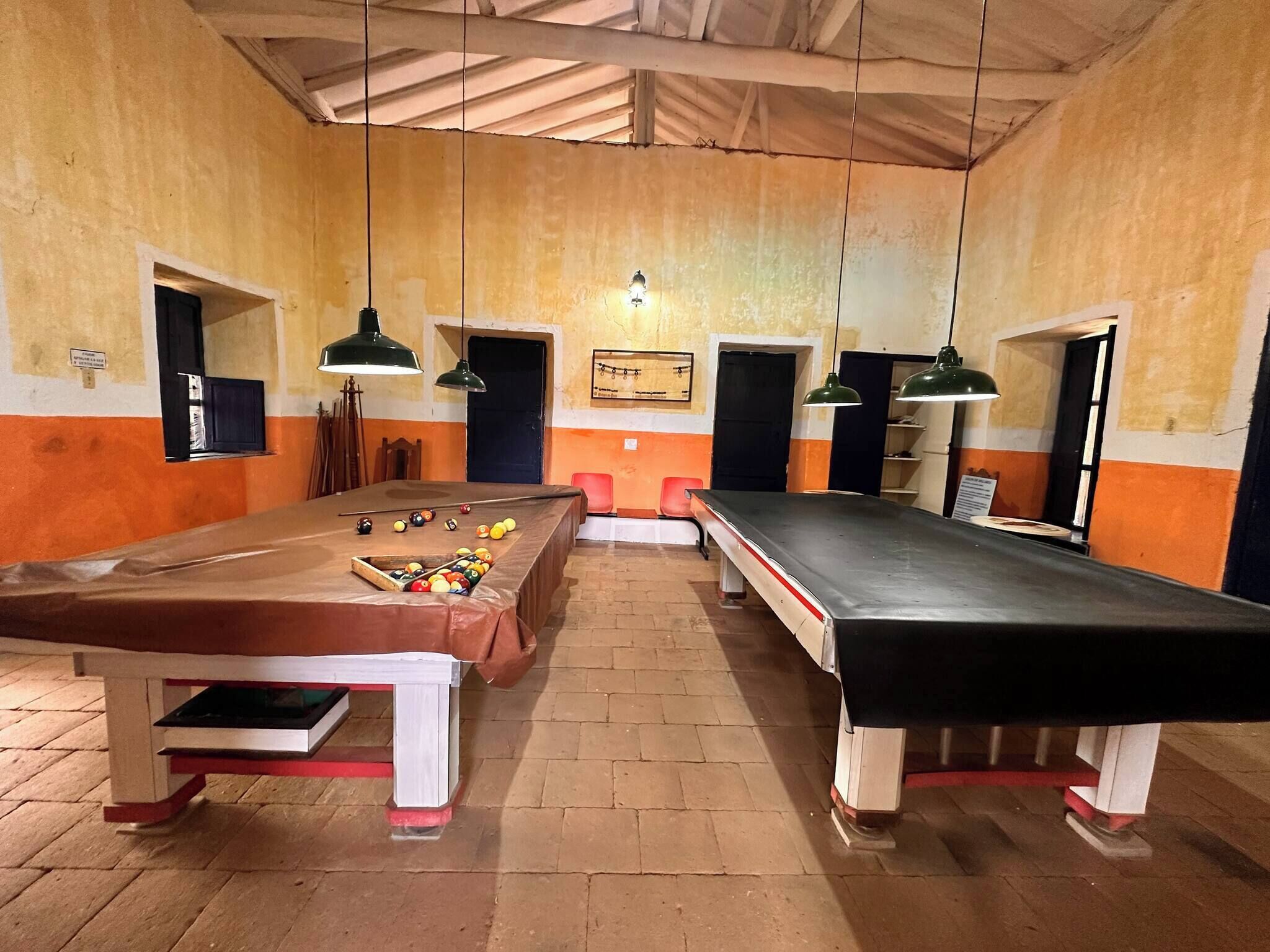 A billiards room with two pool tables, wooden flooring, and hanging lamps.