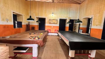 A billiards room with two pool tables, wooden flooring, and hanging lamps.