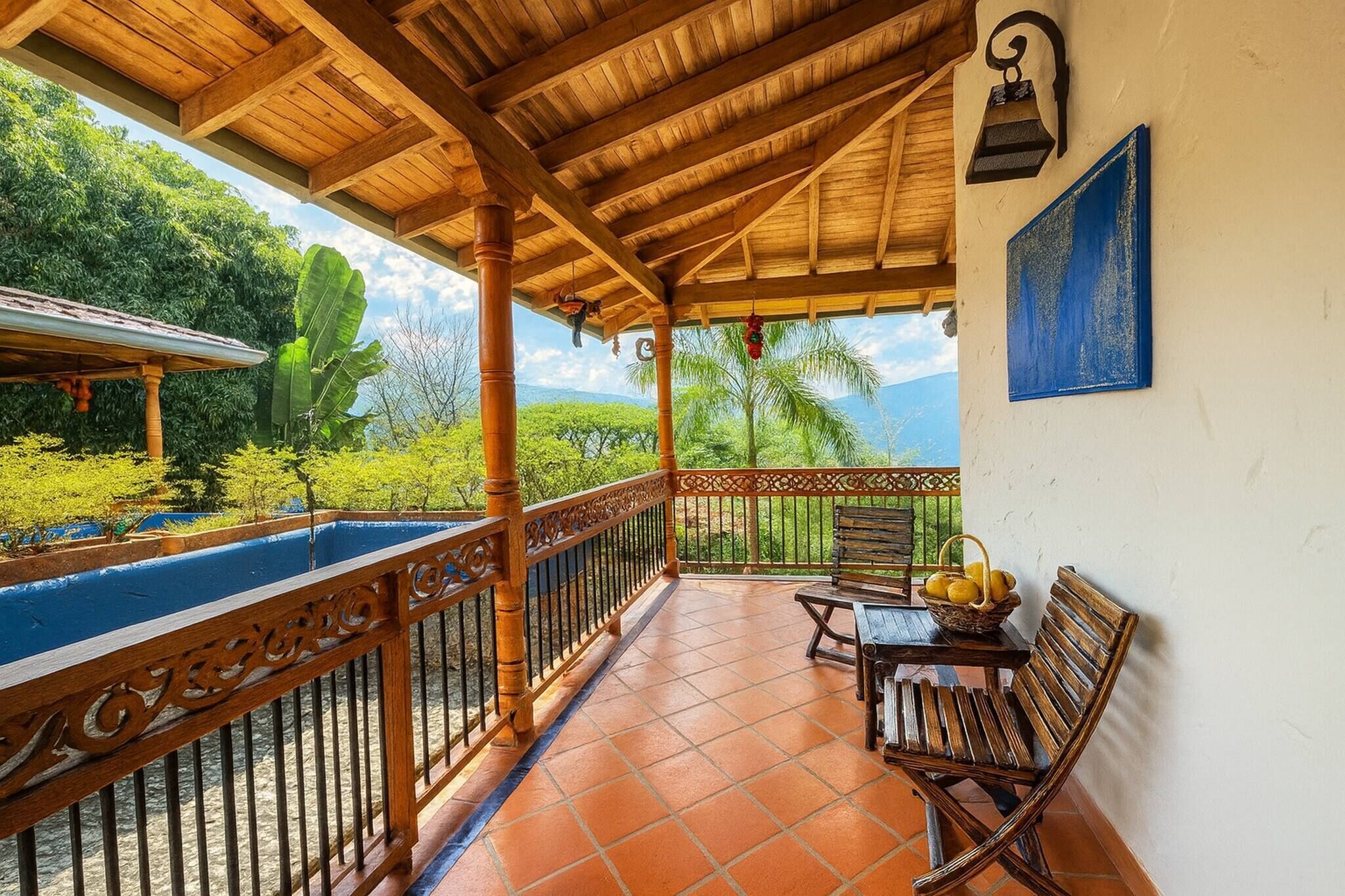 Un espacio al aire libre techado con techo de madera, piso de baldosas, barandilla metálica y vistas a la vegetación y montañas.