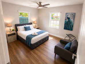 Une chambre à coucher comprenant un lit, des tables de chevet, un ventilateur de plafond, une fenêtre, une chaise et un tableau.