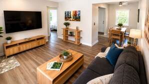 Ein modernes Wohnzimmer mit Flachbildfernseher, Holzmöbeln, Deckenventilator und einem Essbereich mit Tageslicht.